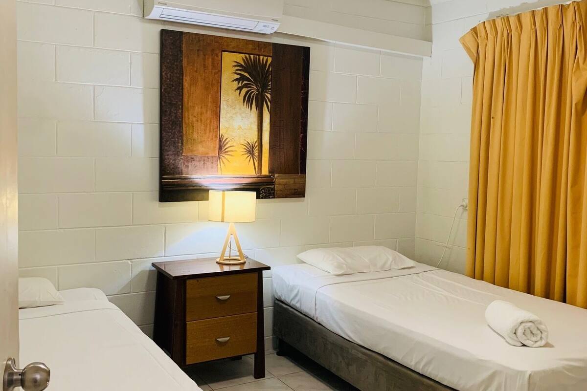 Habitación de hotel con dos camas individuales, una mesita de noche con lámpara, aire acondicionado de pared y un cuadro con palmeras.