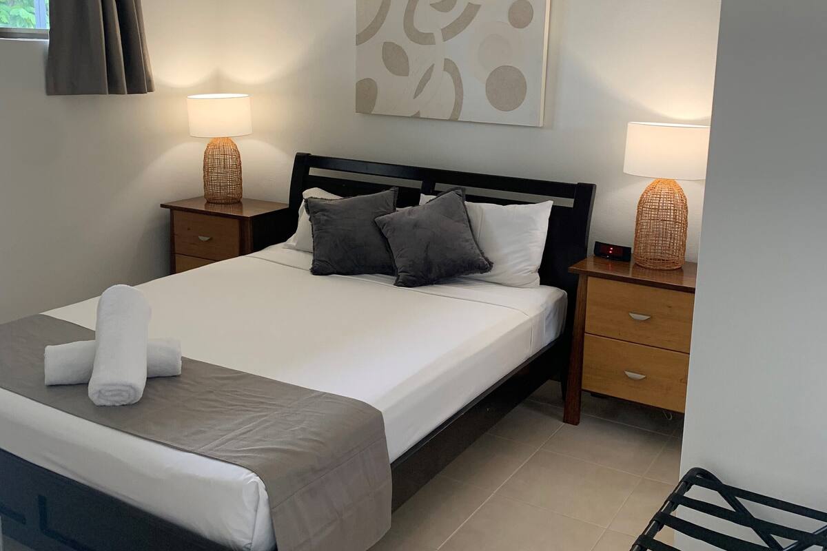 Una cama bien hecha con edredón blanco y gris, dos lámparas de noche, una mesita de noche tejida y una obra de arte en la pared.