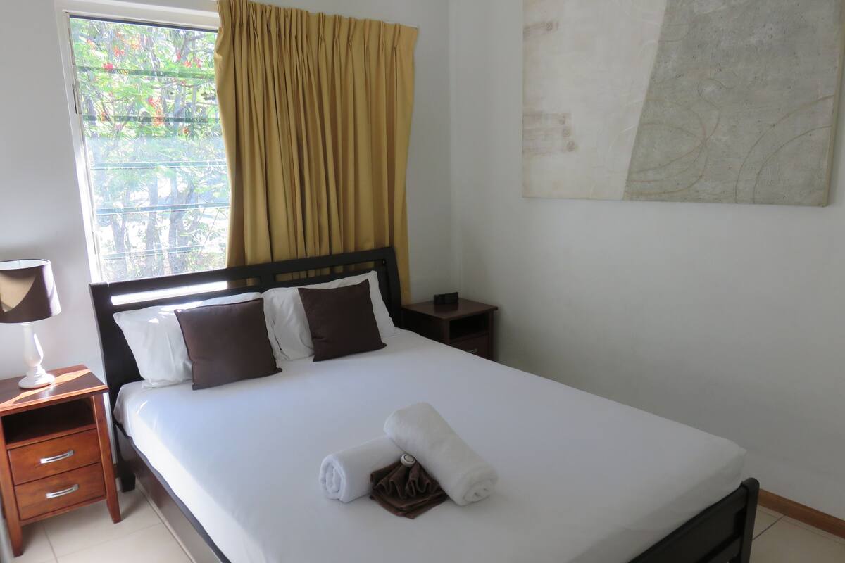 Una cama bien hecha con ropa de cama blanca y marrón, una mesita de noche de madera con una lámpara y una ventana con cortinas amarillas.