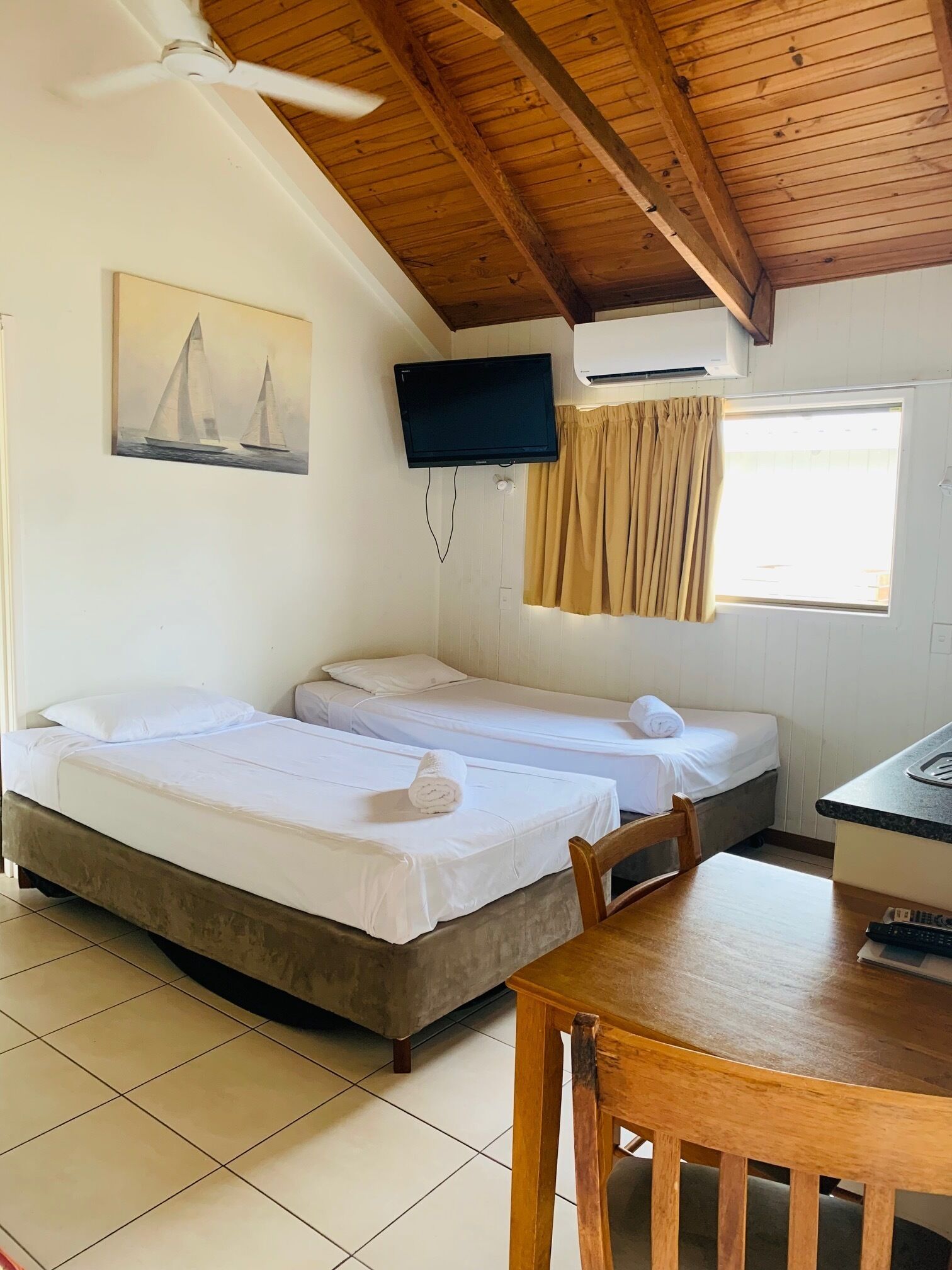 A room with two beds, a ceiling fan, a mounted TV, and a window with curtains.