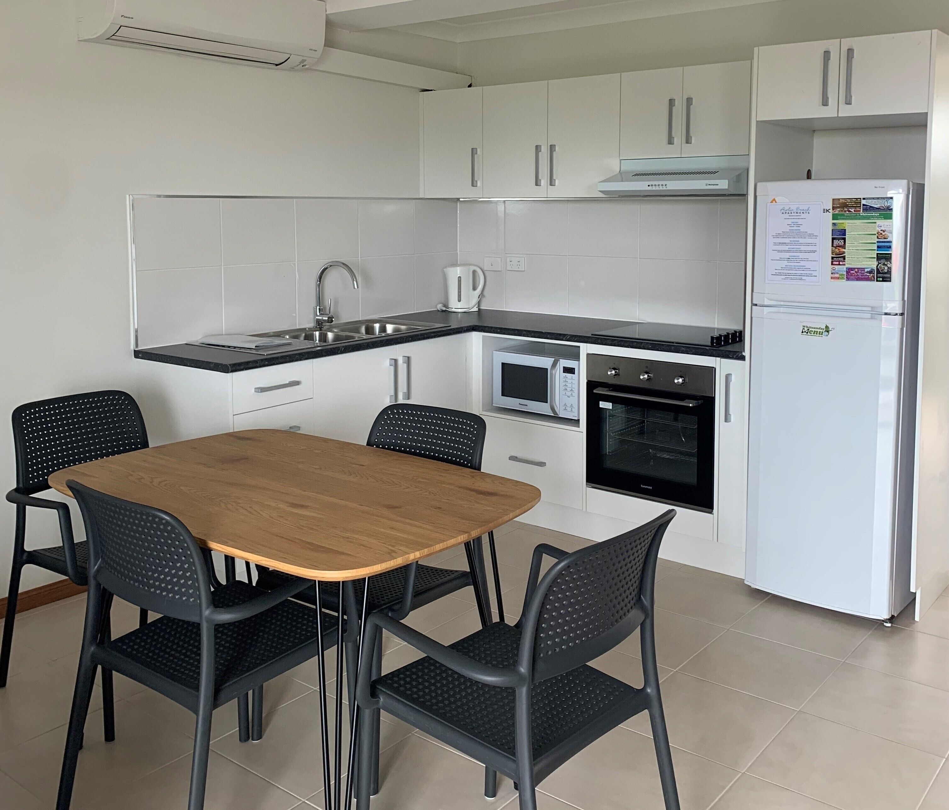 A modern kitchen with white cabinets, a built-in oven, a microwave, and a refrigerator. There is a wooden table with four chairs.