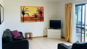 A living room with two black sofas, a small wooden table, a TV on a white cabinet, and a painting of palm trees on the wall. - Airlie Beach Apartments (Airlie Beach)