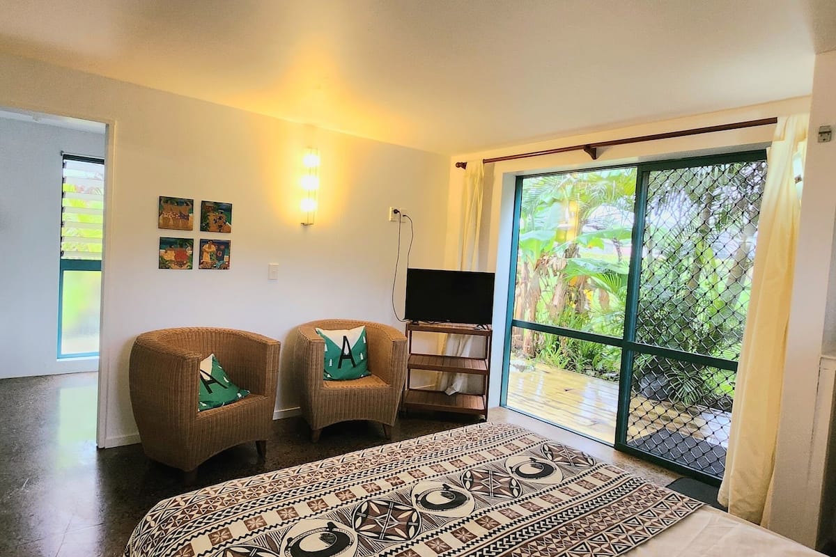 A room with a bed, two chairs, a TV, and a large sliding glass door to a garden.