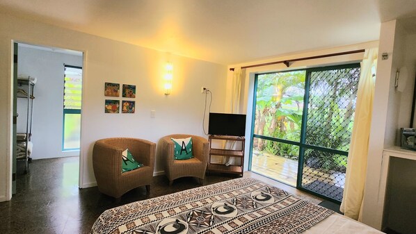Una habitación con una cama, dos sillas, un televisor y un amplio ventanal corredera a un jardín.