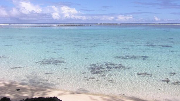 A beach with clear turquoise water, a sandy shore, and distant rocky outcrops. - Aroa Kainga Bungalows (Rarotonga)