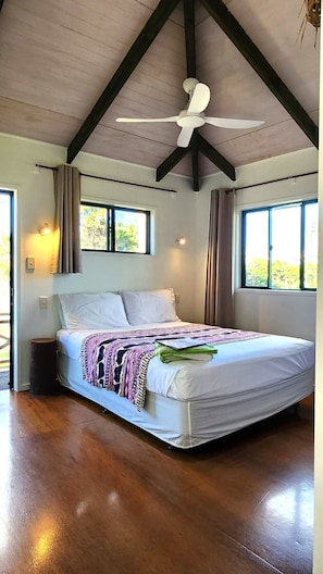 A bedroom with a bed, ceiling fan, windows, and a view of greenery outside. - Aroa Kainga Bungalows (Rarotonga)