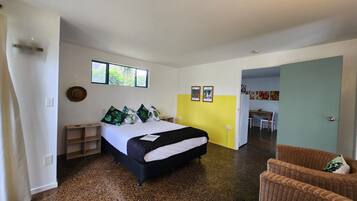 A bedroom with a bed, a nightstand, a chair, and a dining area visible through an open door.