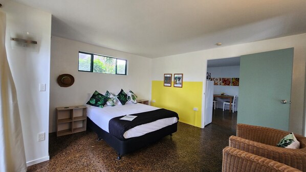 A bedroom with a bed, a nightstand, a chair, and a dining area visible through an open door.