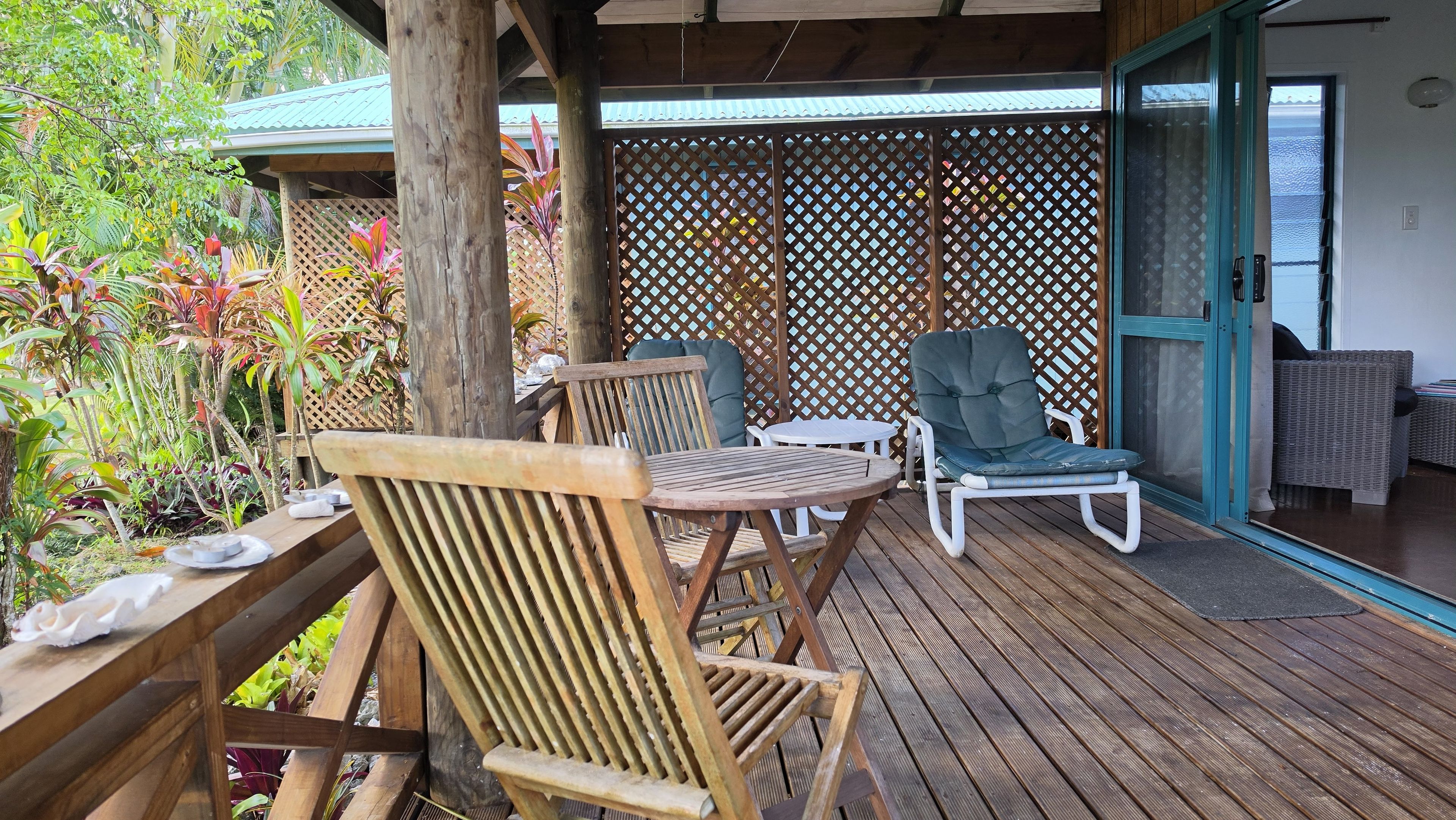 A wooden deck with a table and two chairs, a covered area with a lattice screen, and a view of the sea through a door.