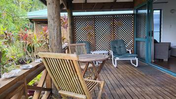 A wooden deck with a table and two chairs, a covered area with a lattice screen, and a view of the sea through a door.