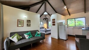 Uma sala de estar com sofá, um espaço de refeições com mesa e cadeiras, uma cozinha com geladeira e uma janela grande com vista para o verde.