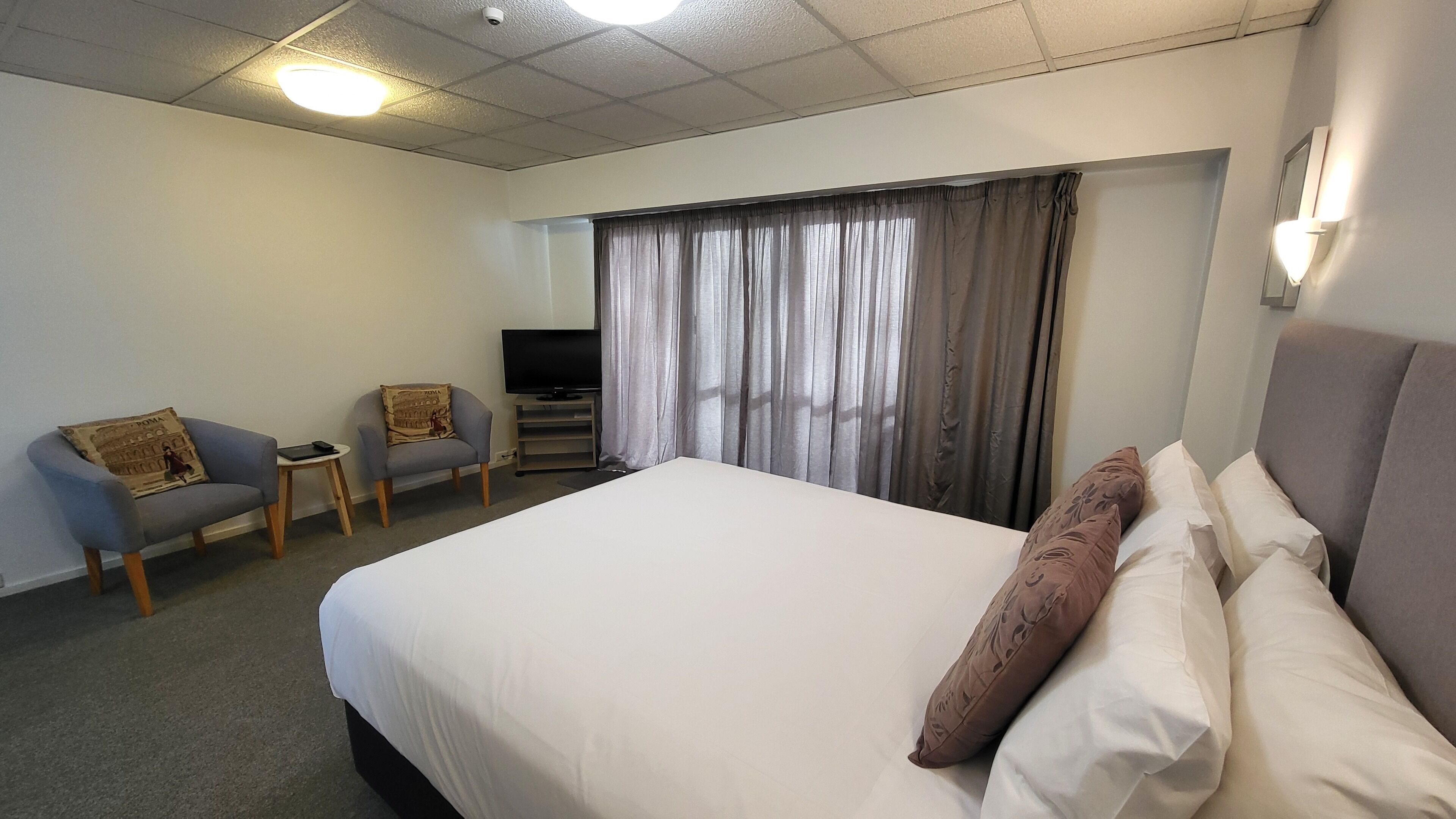 A hotel room with a large bed, two armchairs, a small table, a TV, and a window with curtains.