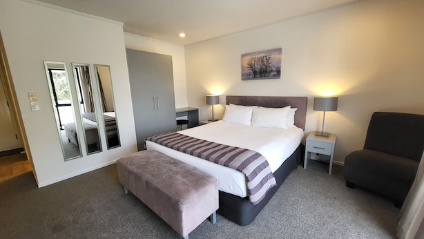 A neatly arranged bedroom with a large bed, bedside lamps, a chair, and a mirror. - Manukau Motor Lodge (Auckland)