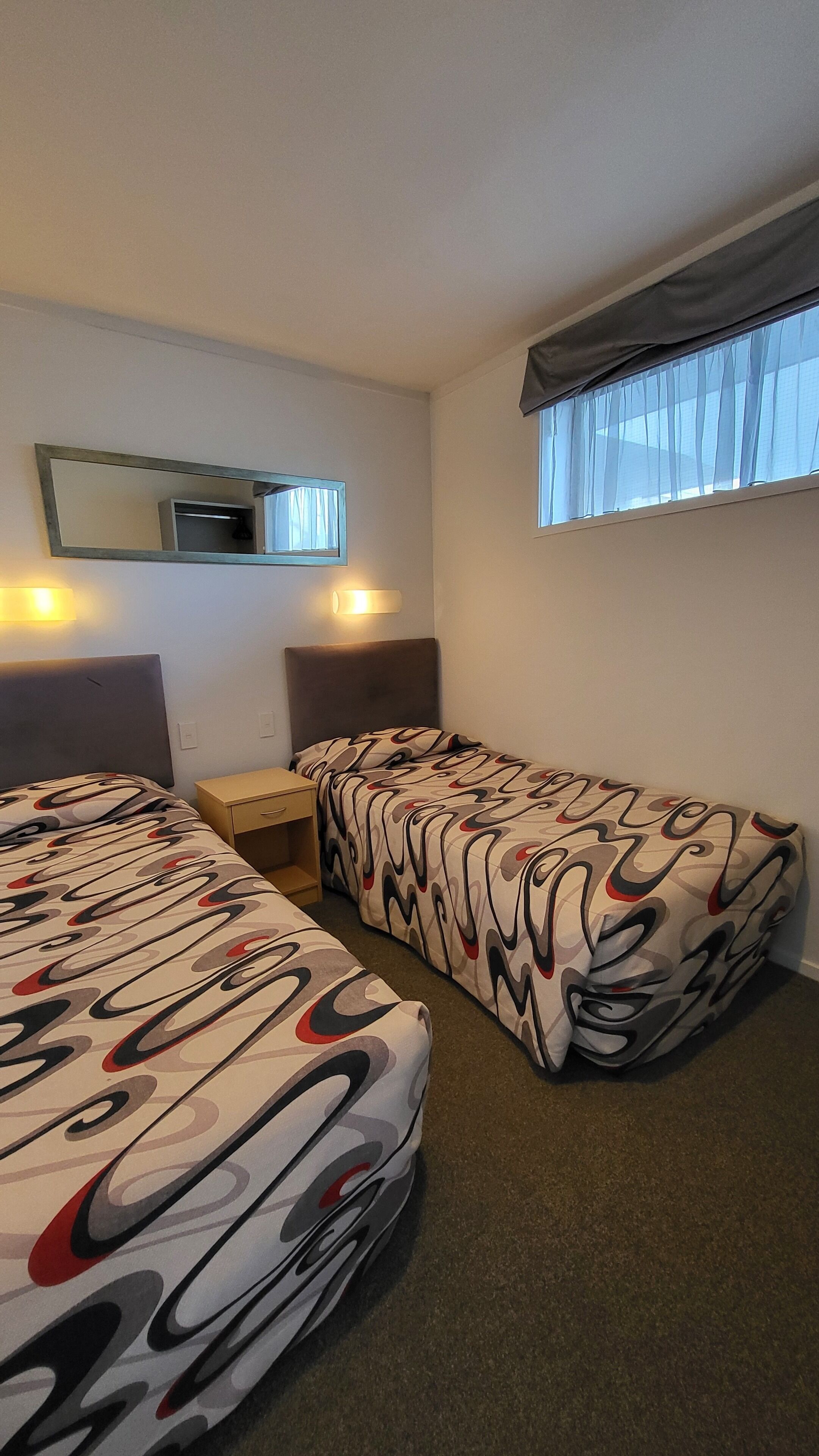 A hotel room with two beds, a small table, a mirror, and a window with curtains.