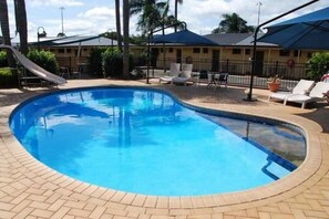 A swimming pool with a slide, surrounded by lounge chairs and umbrellas, with a building in the background. - James Street Motor Inn (South Toowoomba)