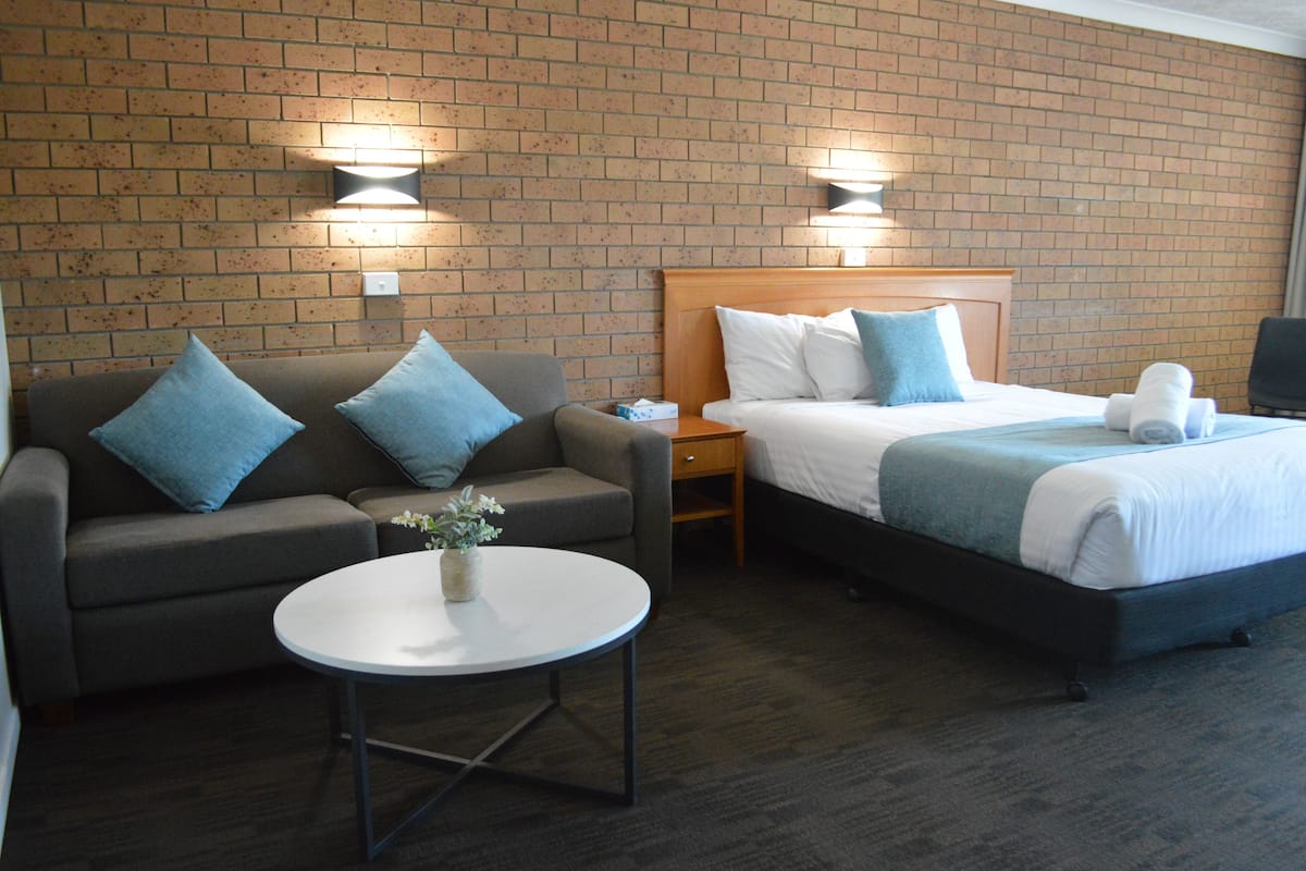 A hotel room with a brick wall, a bed with white and blue bedding, a grey sofa with blue cushions, a small table with a plant, and two wall-mounted lights.