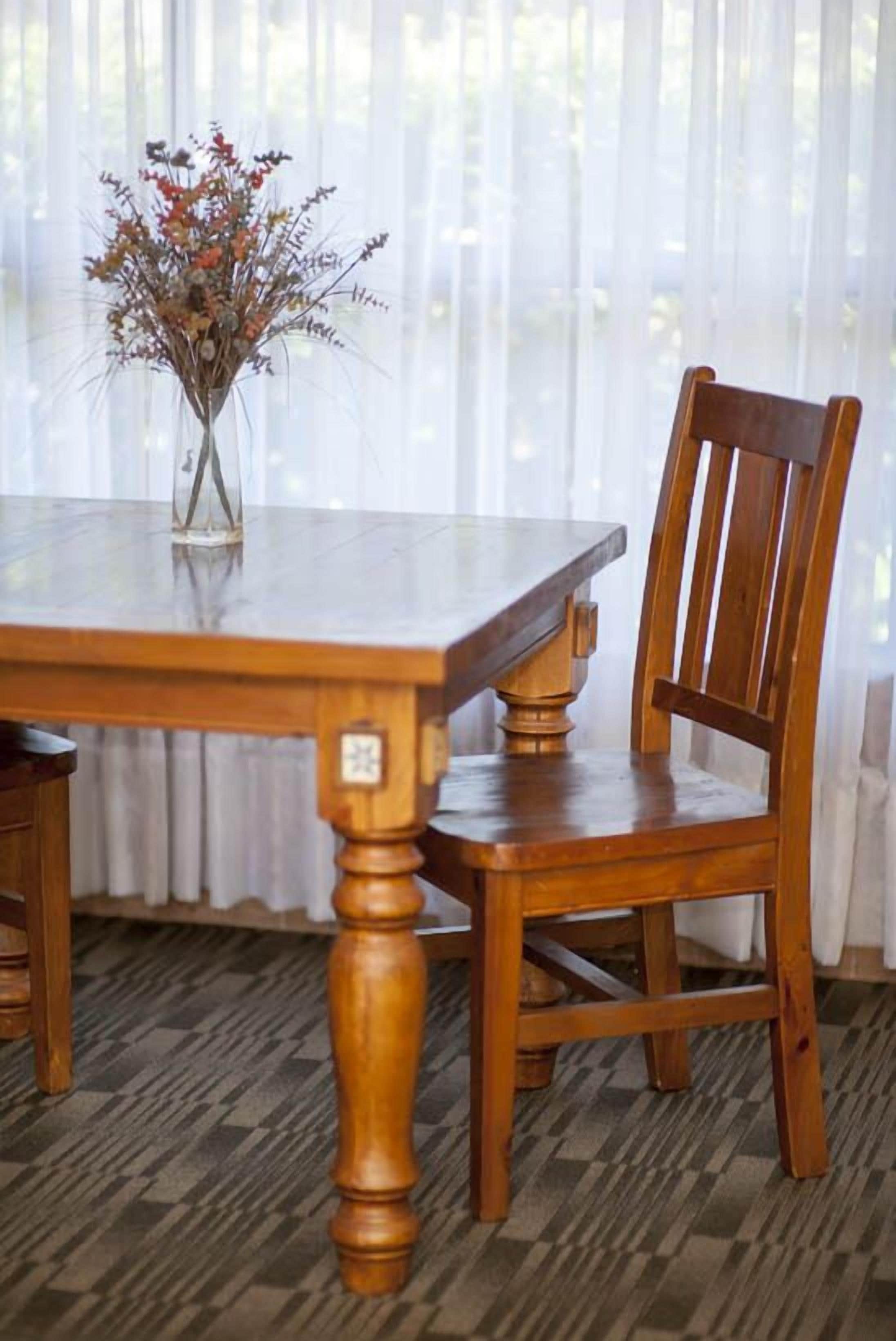 Una mesa de comedor de madera con un jarrón de flores secas, ubicada frente a una ventana con cortinas translúcidas.