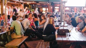 Un bar concurrido con mesas y bancos de madera, clientes sentados y de pie, y un letrero verde arriba.
