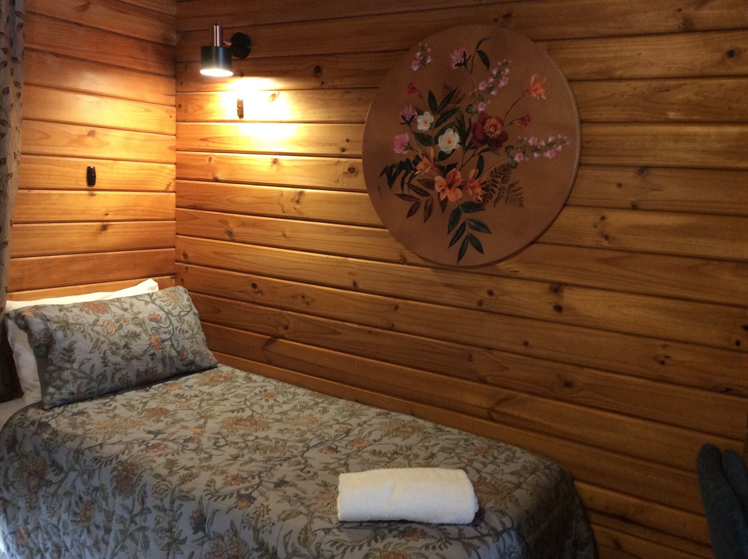 Una habitación tipo cabaña de madera con una cama, una lámpara fijada a la pared y una obra de arte floral.