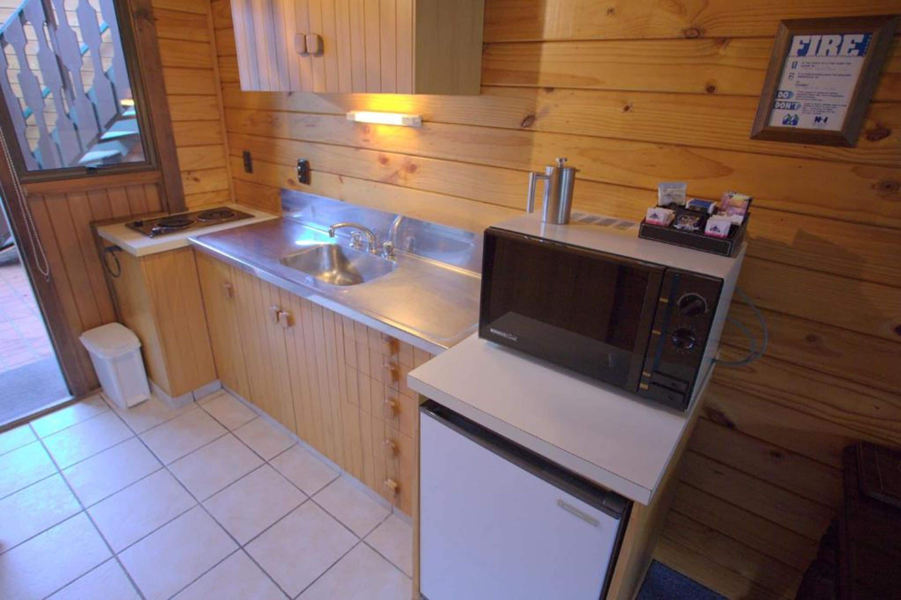 A compact kitchen with wooden walls, a stainless steel sink, a microwave, and a small refrigerator.