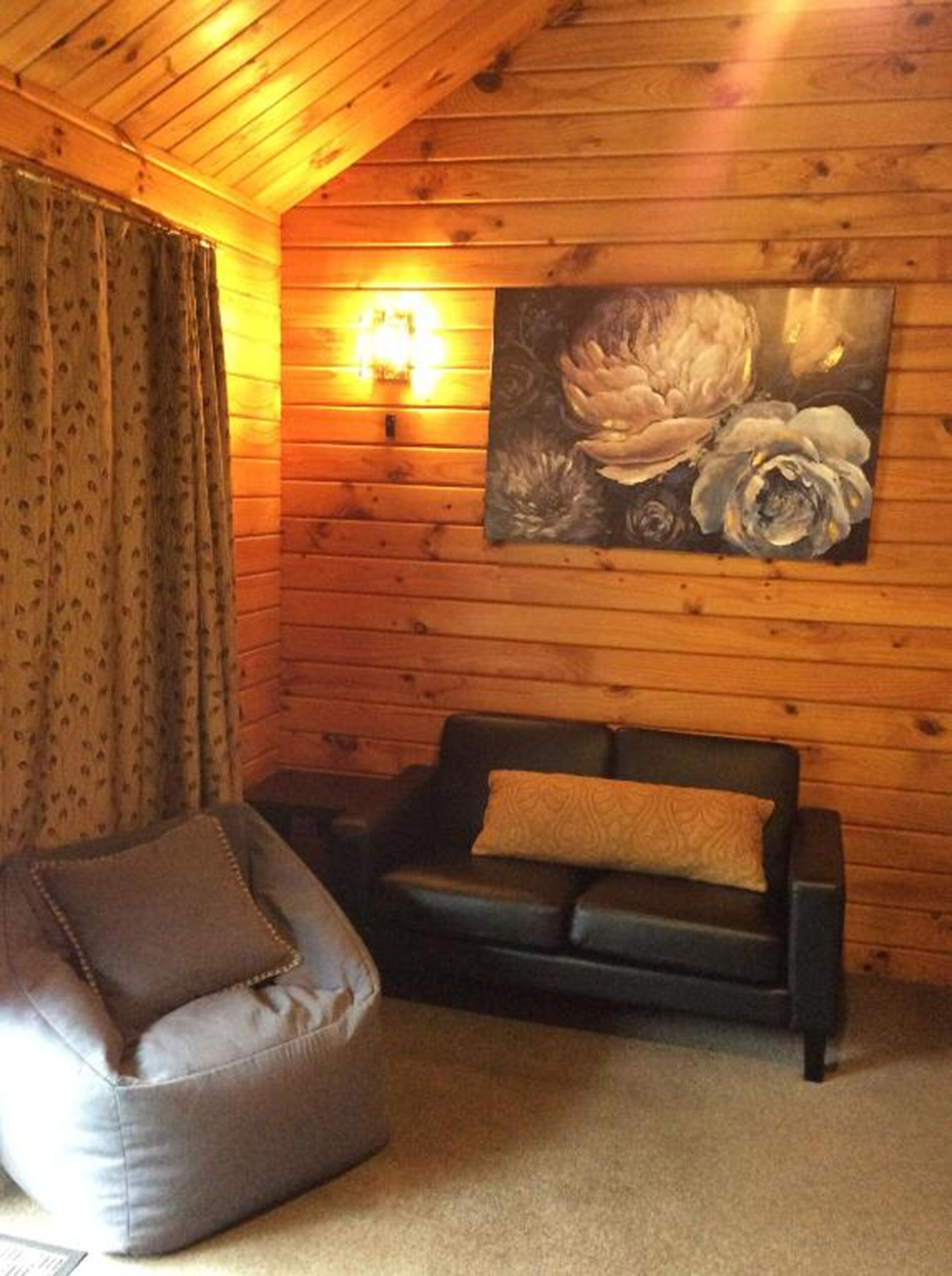A cozy room with a wooden interior, a black sofa with a cushion, a bean bag chair, and a floral painting on the wall.