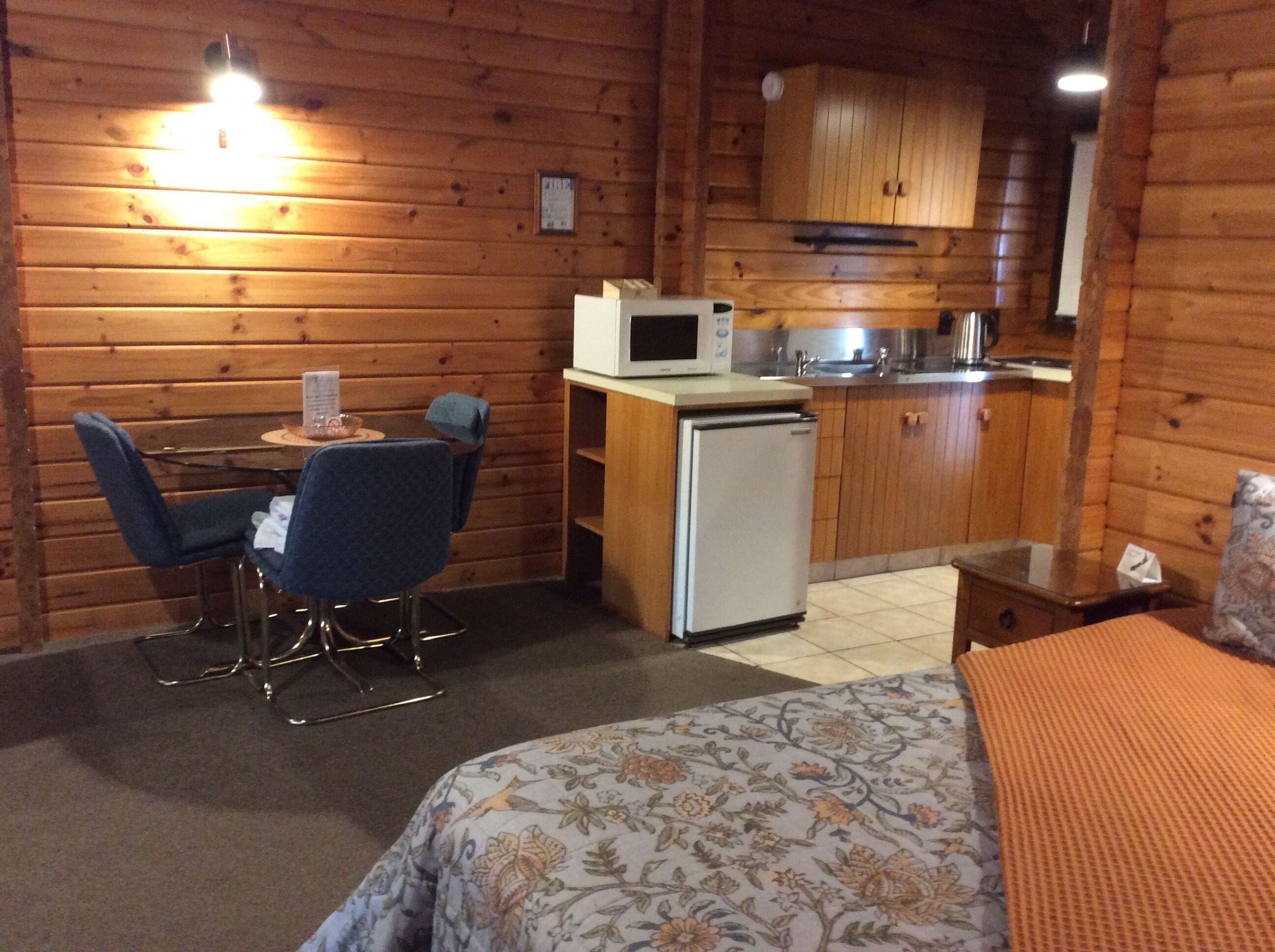 Una habitación compacta con cama, una kitchenette pequeña, una zona de comedor con mesa y sillas, y paredes de madera.
