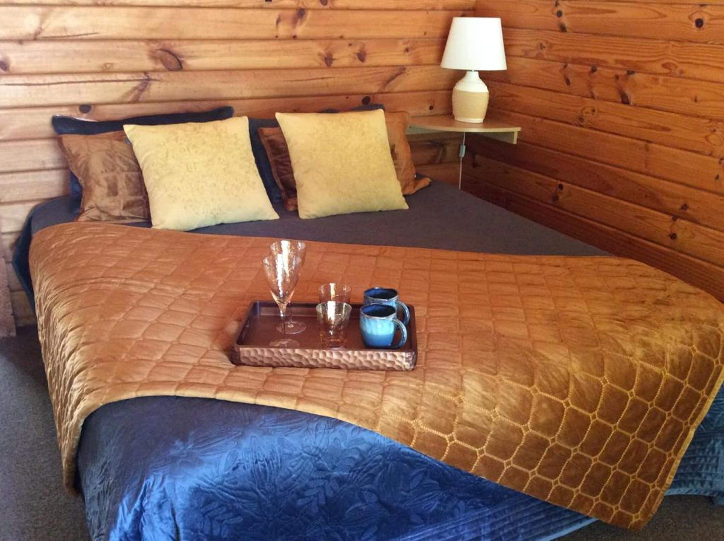A wooden cabin room with a bed, two pillows, a nightstand with a lamp, and a tray with a glass and mugs.