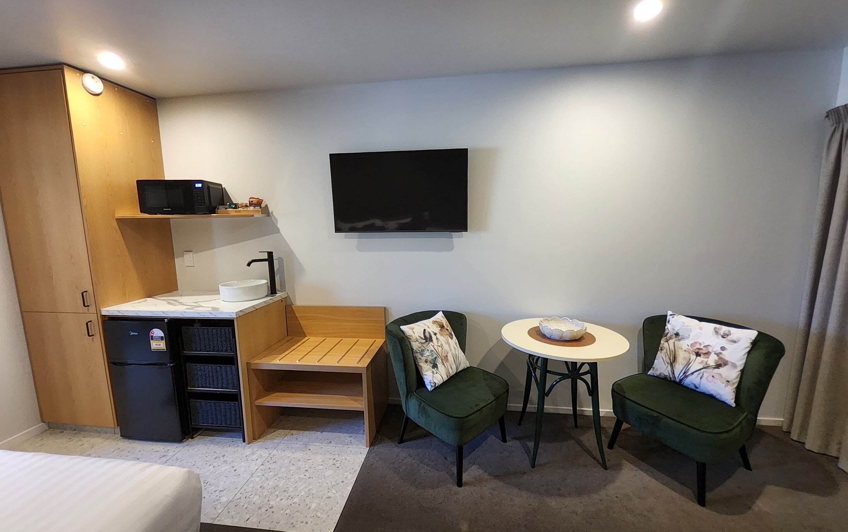 A compact hotel room with a bed, a small kitchenette, a TV, two green chairs, a round table, and a wooden bench.