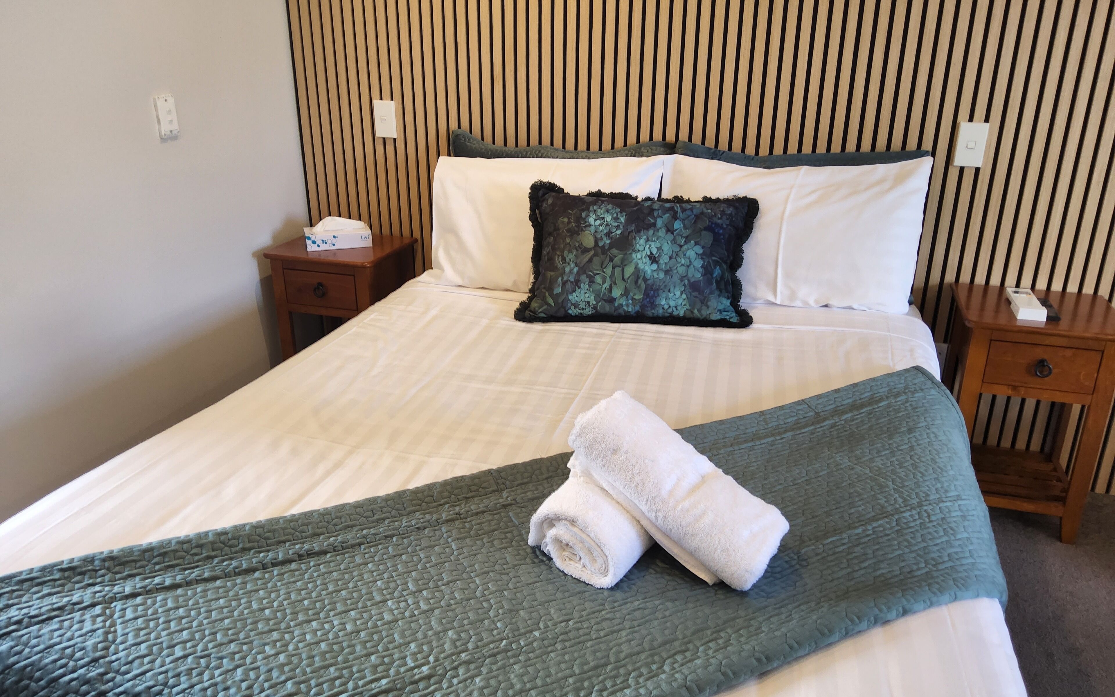 A neatly made bed with a green quilt, white pillows, and a rolled white towel on the bed. There are two wooden nightstands with a box on one and a phone on the other.