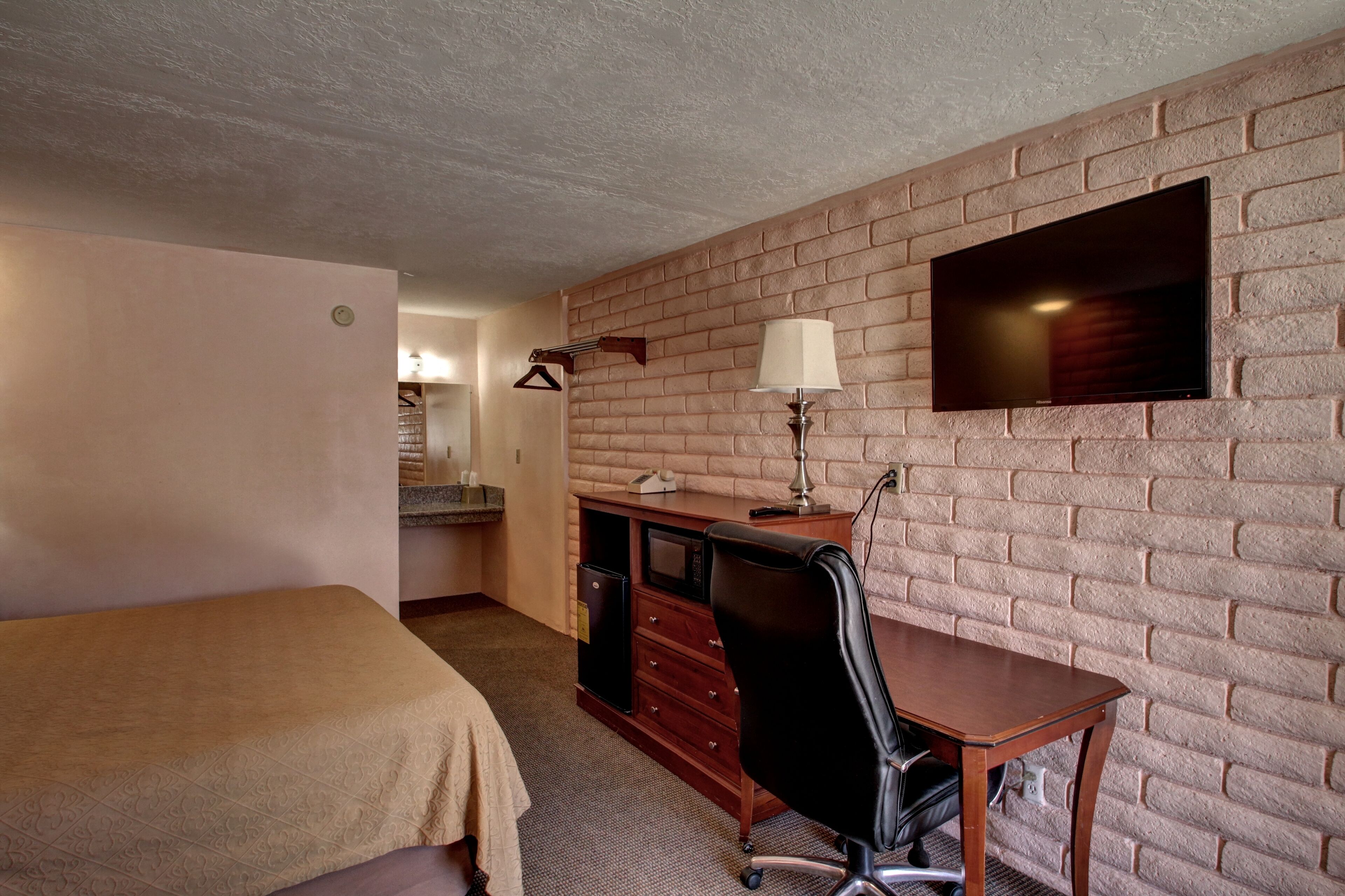 Habitación de hotel con cama, escritorio con silla, televisor montado en pared de ladrillo y lámpara.