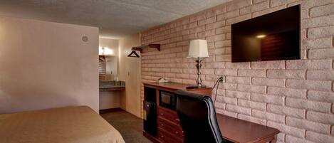 A hotel room with a bed, a desk with a chair, a TV mounted on a brick wall, and a lamp.