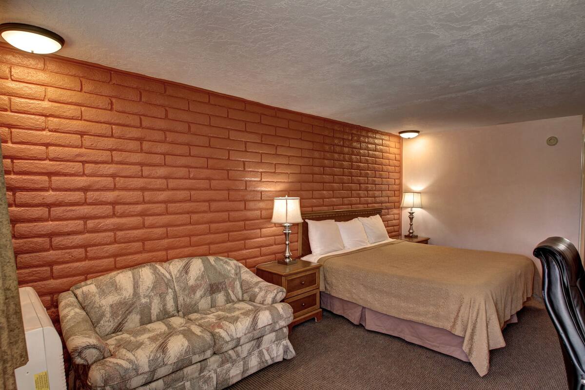 A hotel room with a brick-patterned wall, a bed with a beige bedspread, a patterned armchair, and two bedside lamps.