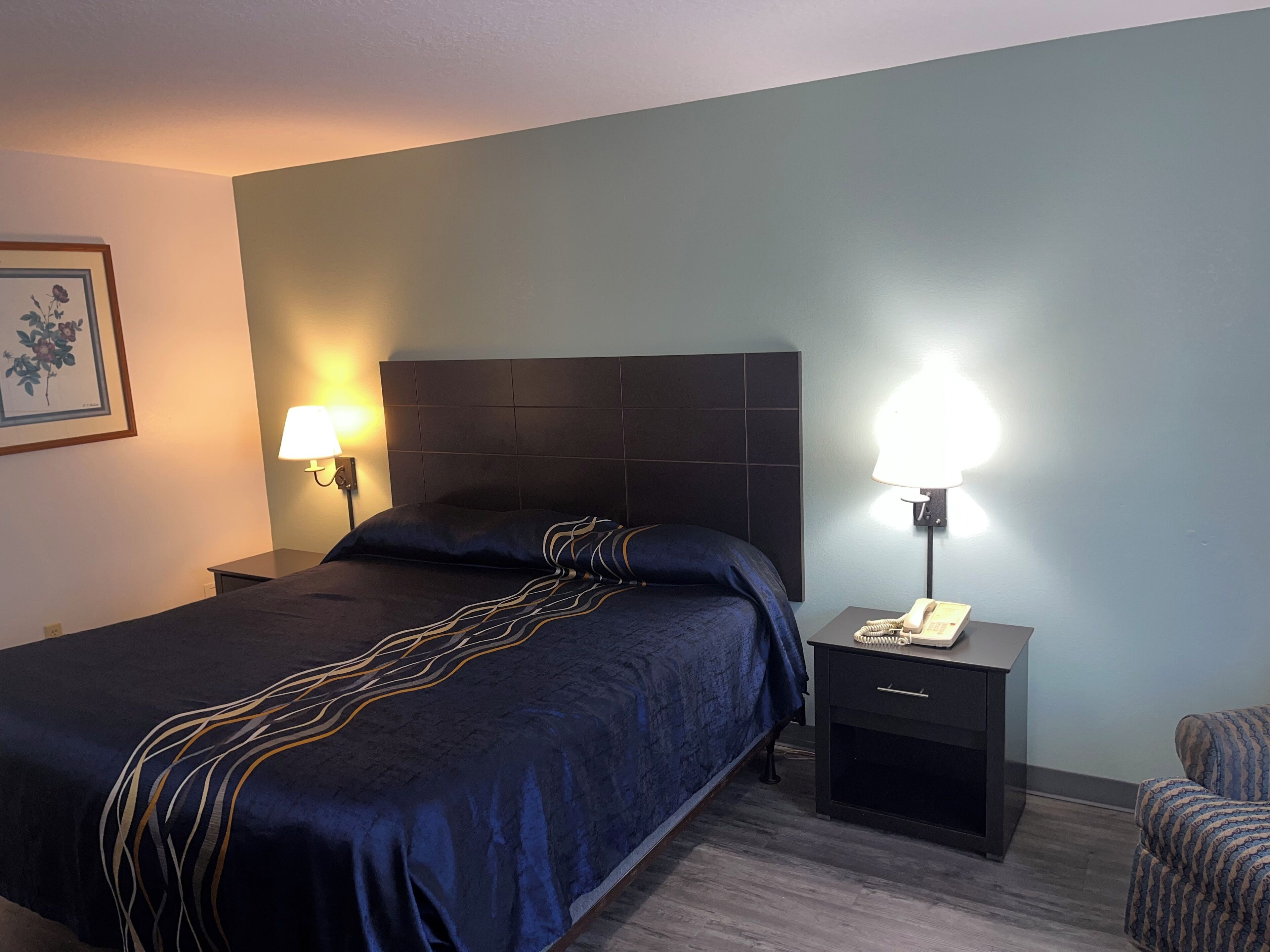 A hotel room with a large bed, two bedside tables, a nightstand, a chair, and a framed picture on the wall.