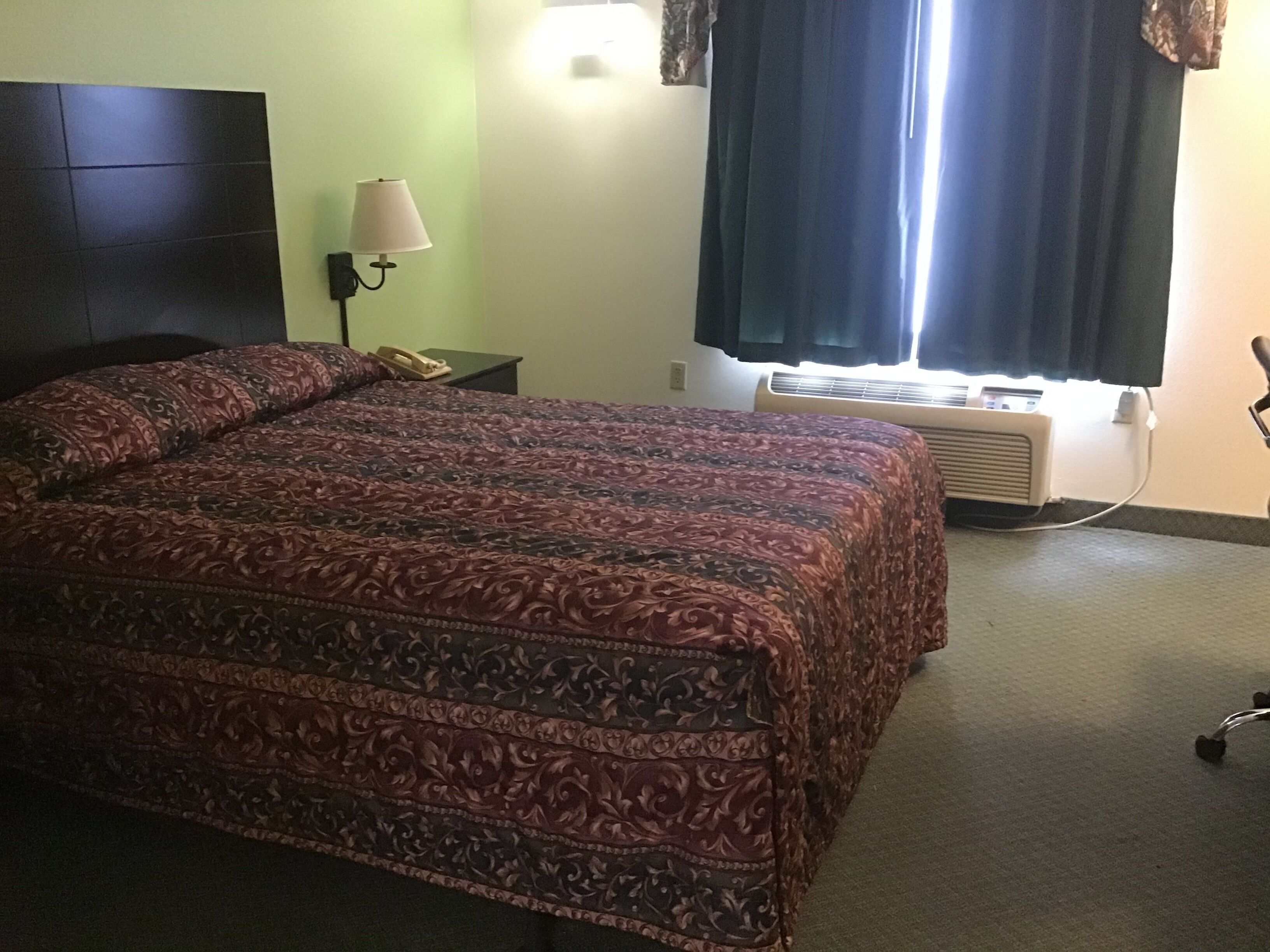 A hotel room with a large bed, a bedside lamp, a telephone, an air conditioning unit, and a window with curtains.