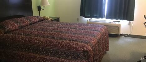 A hotel room with a large bed, a bedside lamp, a telephone, an air conditioning unit, and a window with curtains.