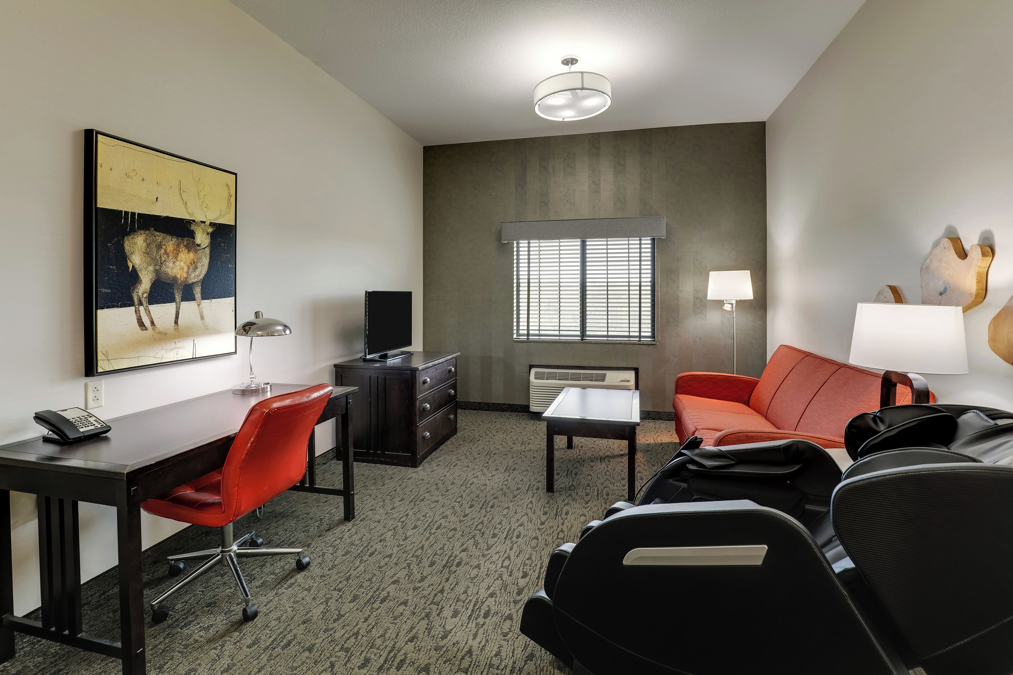 A hotel room with a desk, red office chair, black leather sofa, and a large painting of a deer.