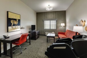 Una habitación de hotel con escritorio, silla de oficina roja, sofá de cuero negro y un cuadro grande de un ciervo.