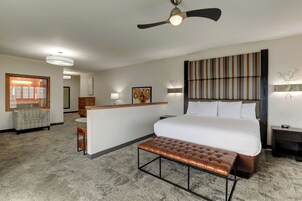 Habitación de hotel con una cama grande, un escritorio, un banco, un ventilador de techo y vistas al pasillo.