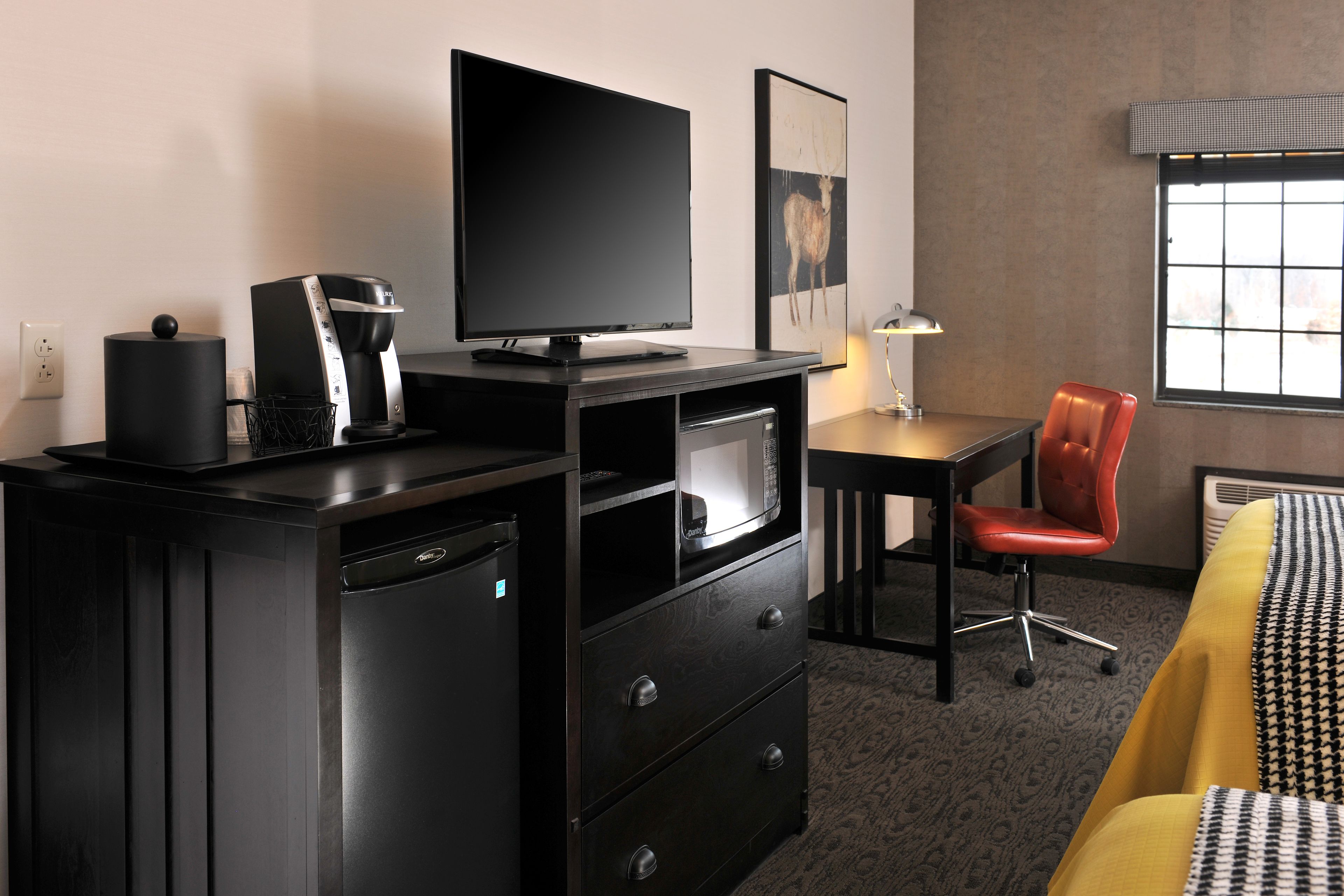 A hotel room with a black TV, a coffee maker, a microwave, a desk, a red chair, and a bed with a patterned bedspread.