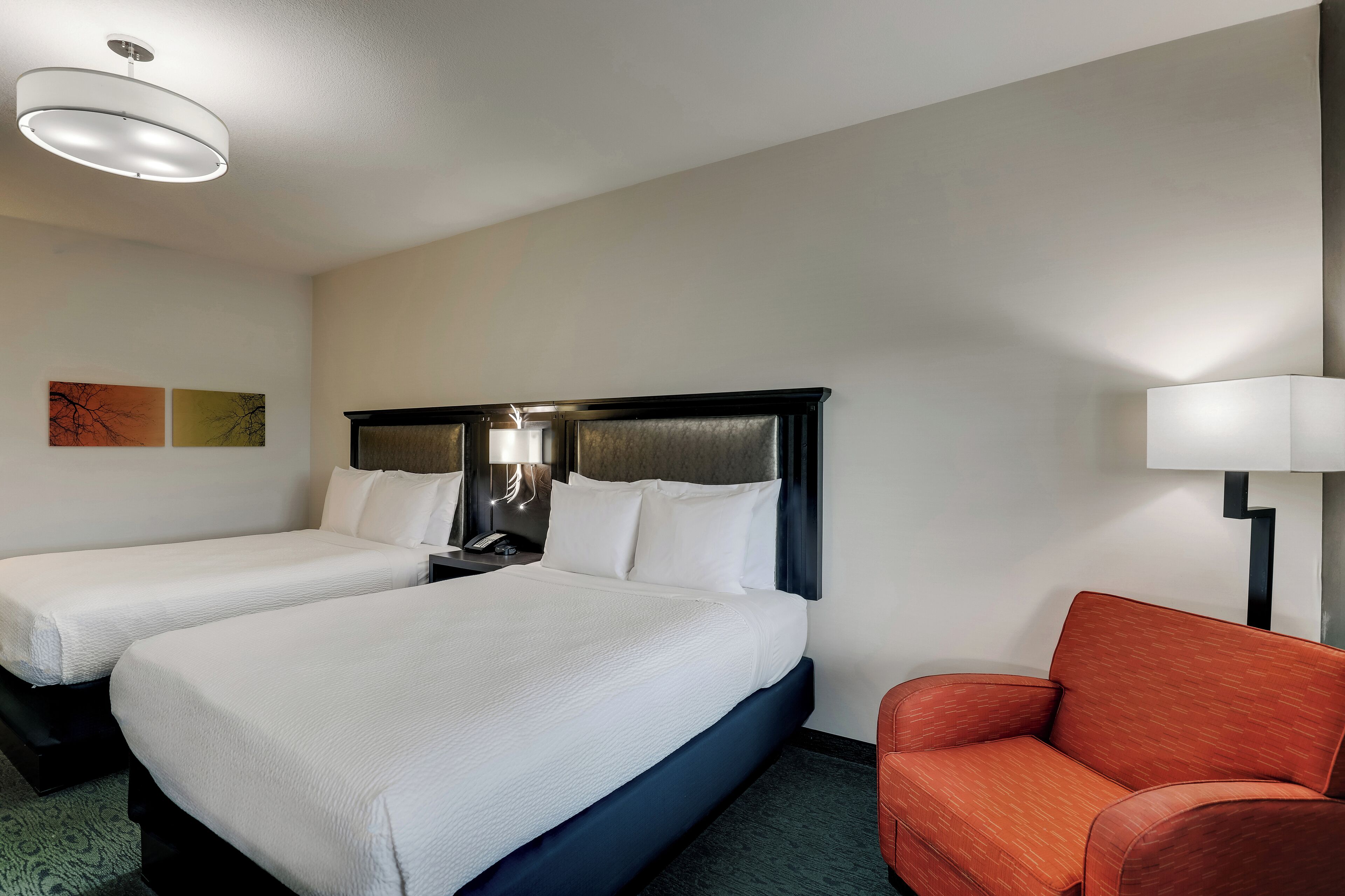 Une chambre d’hôtel avec deux lits, une table de chevet avec une lampe, un fauteuil rouge et un luminaire au plafond.