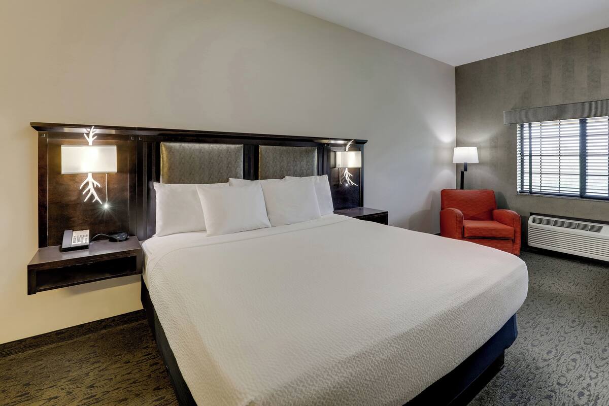 Une chambre d’hôtel avec un grand lit, une table de chevet avec un téléphone et une lampe, une chaise et une fenêtre avec des stores.