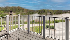 Een houten terras met een reling, met uitzicht op een rivier, omringd door bomen en een gebouw in de verte.