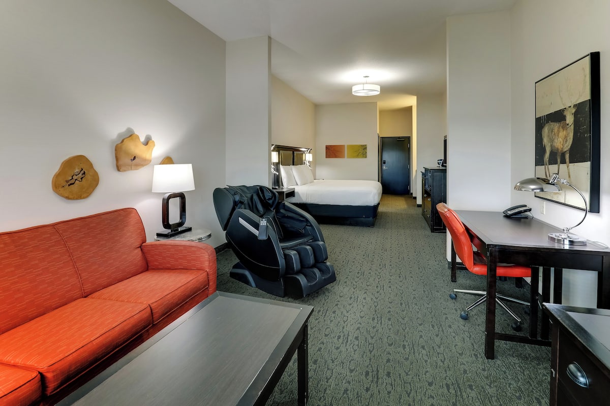 Une chambre d’hôtel avec un canapé rouge, un fauteuil massant, un bureau avec une lampe, un lit et une œuvre d’art accrochée au mur.