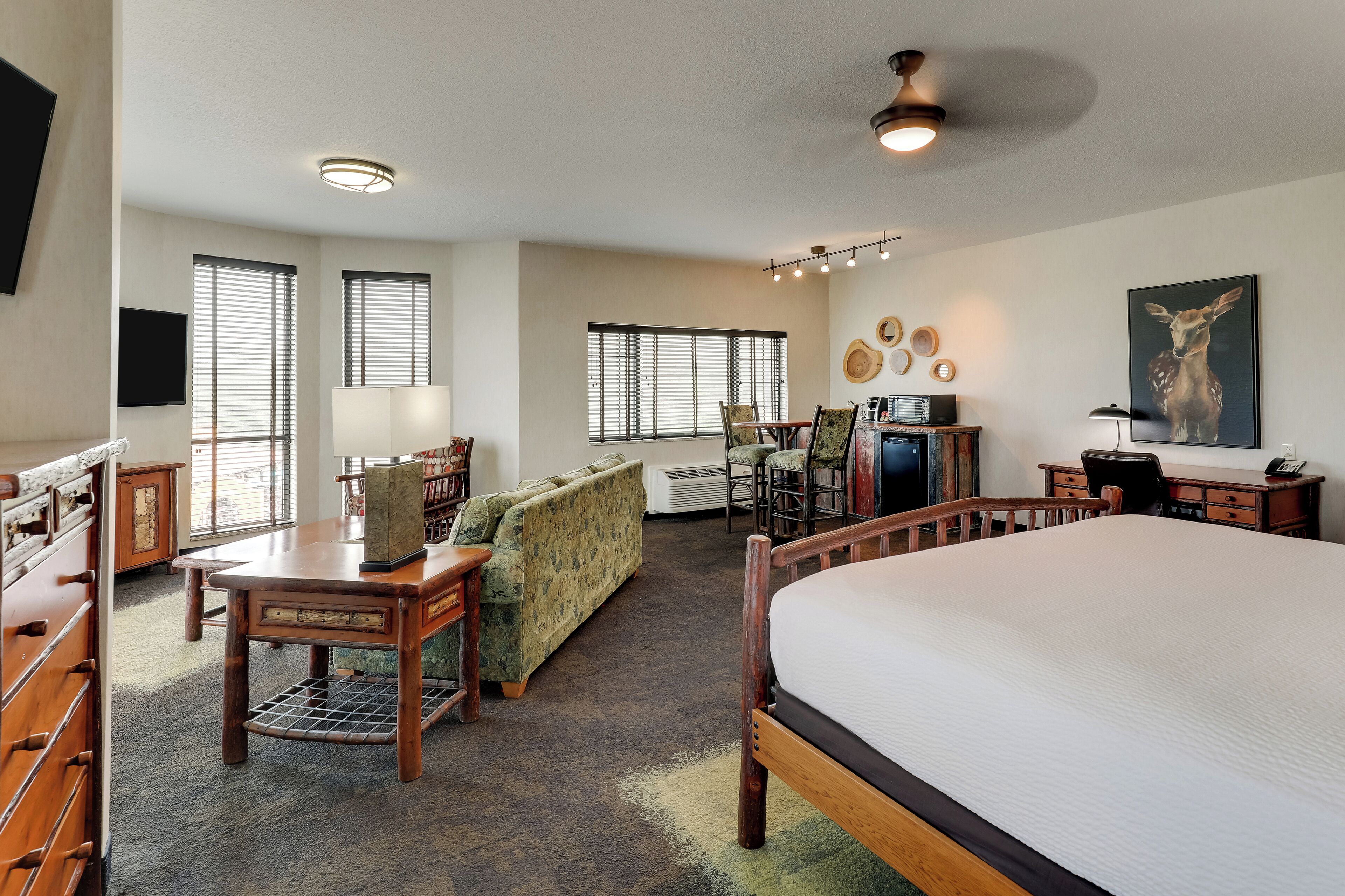 A spacious hotel room with a large bed, a wooden desk, a sofa, and a dining table. There are multiple windows providing natural light, and a ceiling light fixture.
