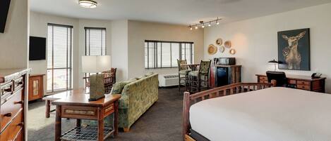Amplia habitación de hotel con una cama grande, un escritorio de madera, un sofá y una mesa de comedor. Varias ventanas ofrecen luz natural, y hay una lámpara de techo.