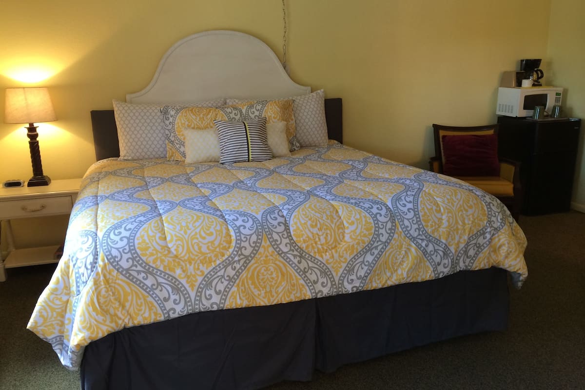A bed with a patterned bedspread, a bedside lamp, a nightstand, a chair, and a microwave on a table.