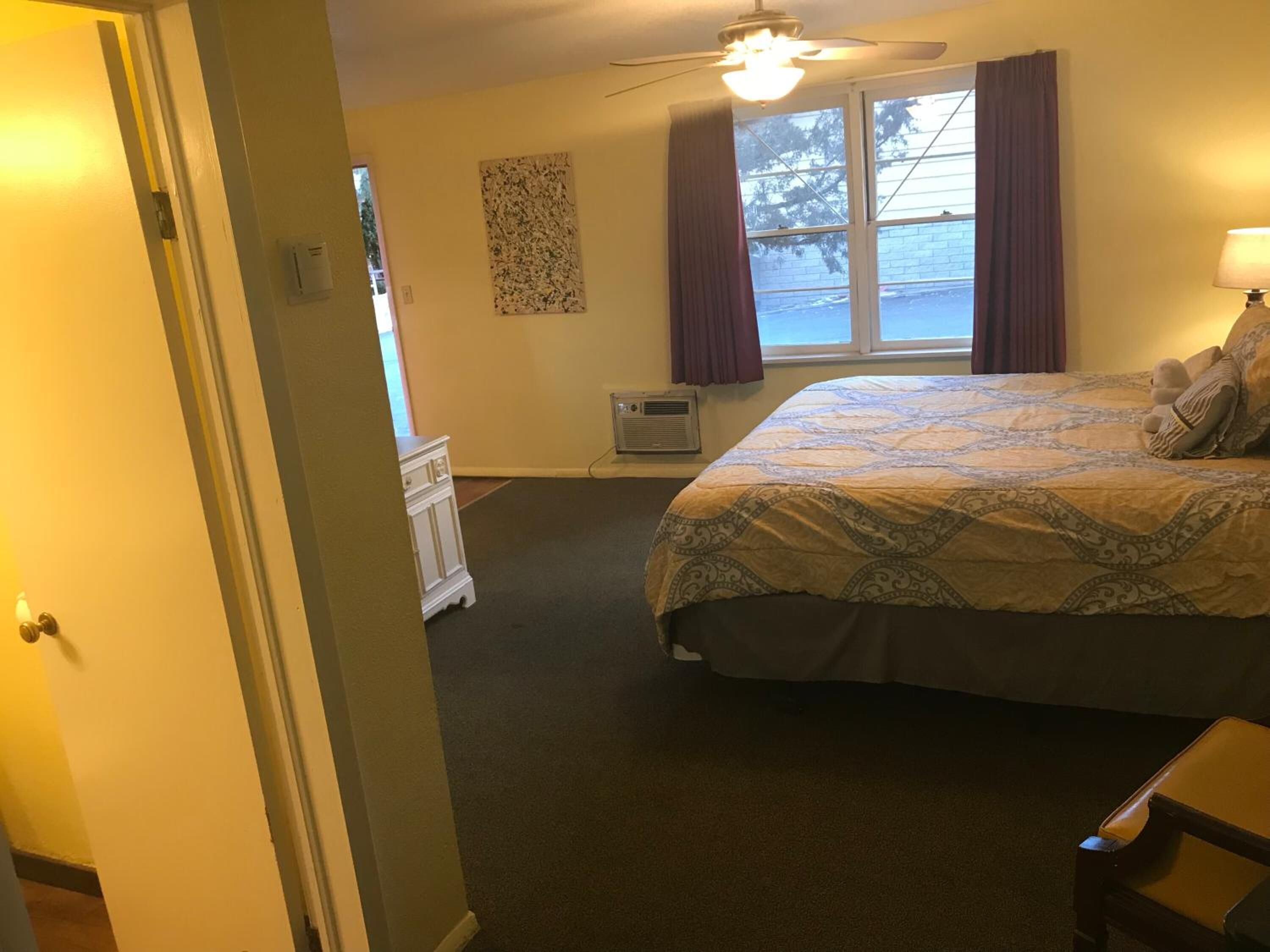 Quarto de hotel com uma cama grande, ventoinha no teto, janela com cortinas e um pequeno ar condicionado.