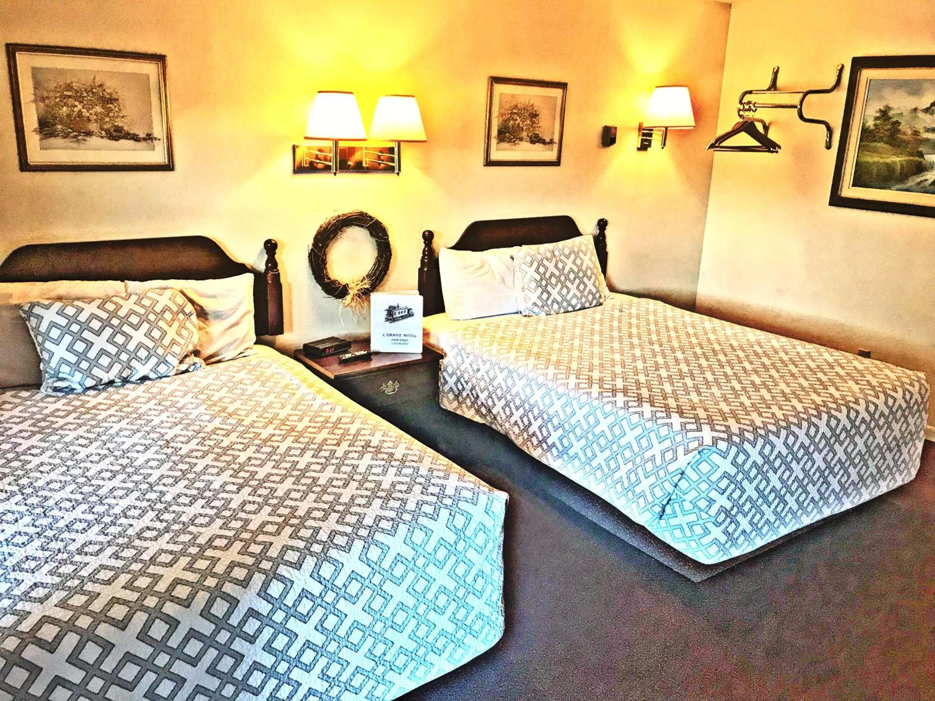 Una habitación de hotel con dos camas individuales, cada una con ropa de cama estampada, una mesita de noche con lámpara y cuadros en las paredes.