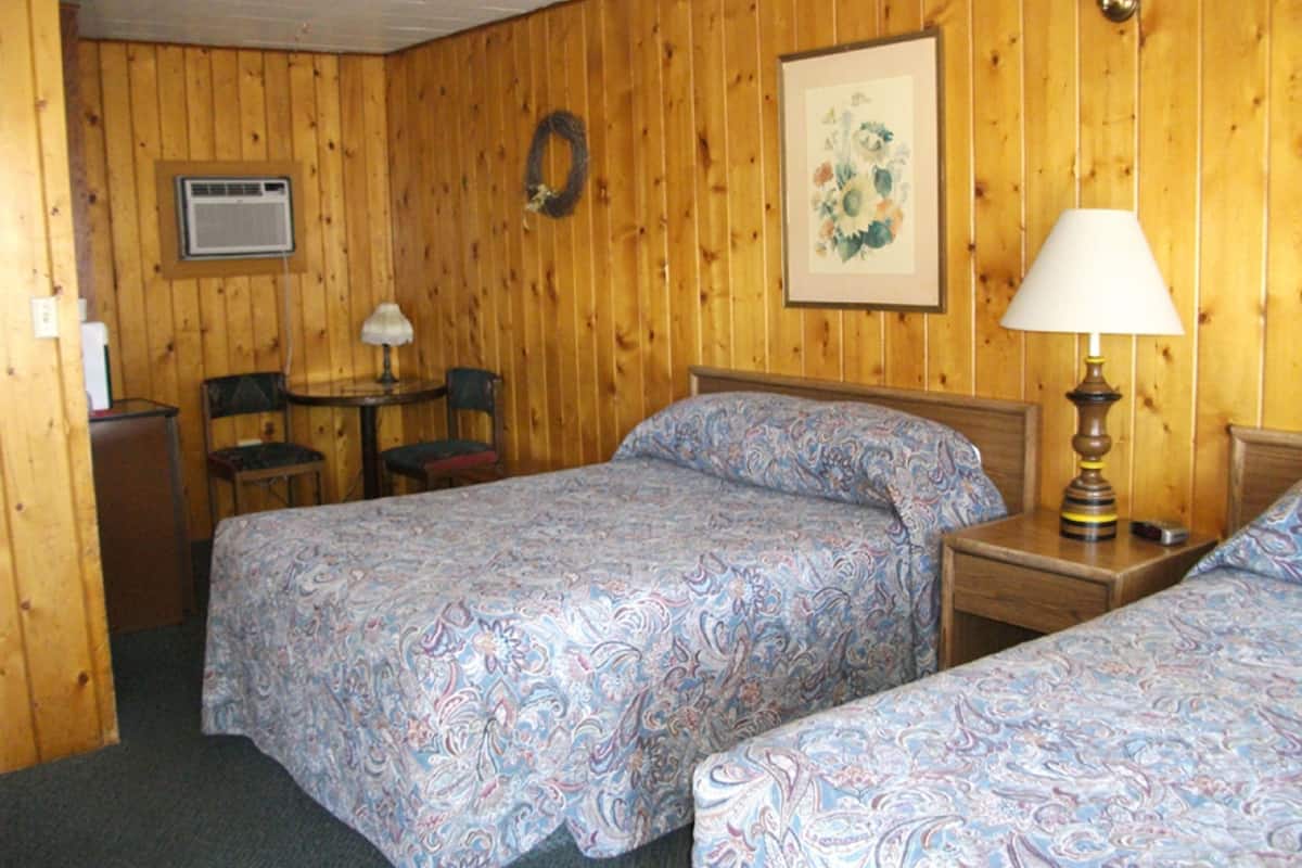 A room with two beds, a small table with a chair, a lamp, and a wall-mounted air conditioner.