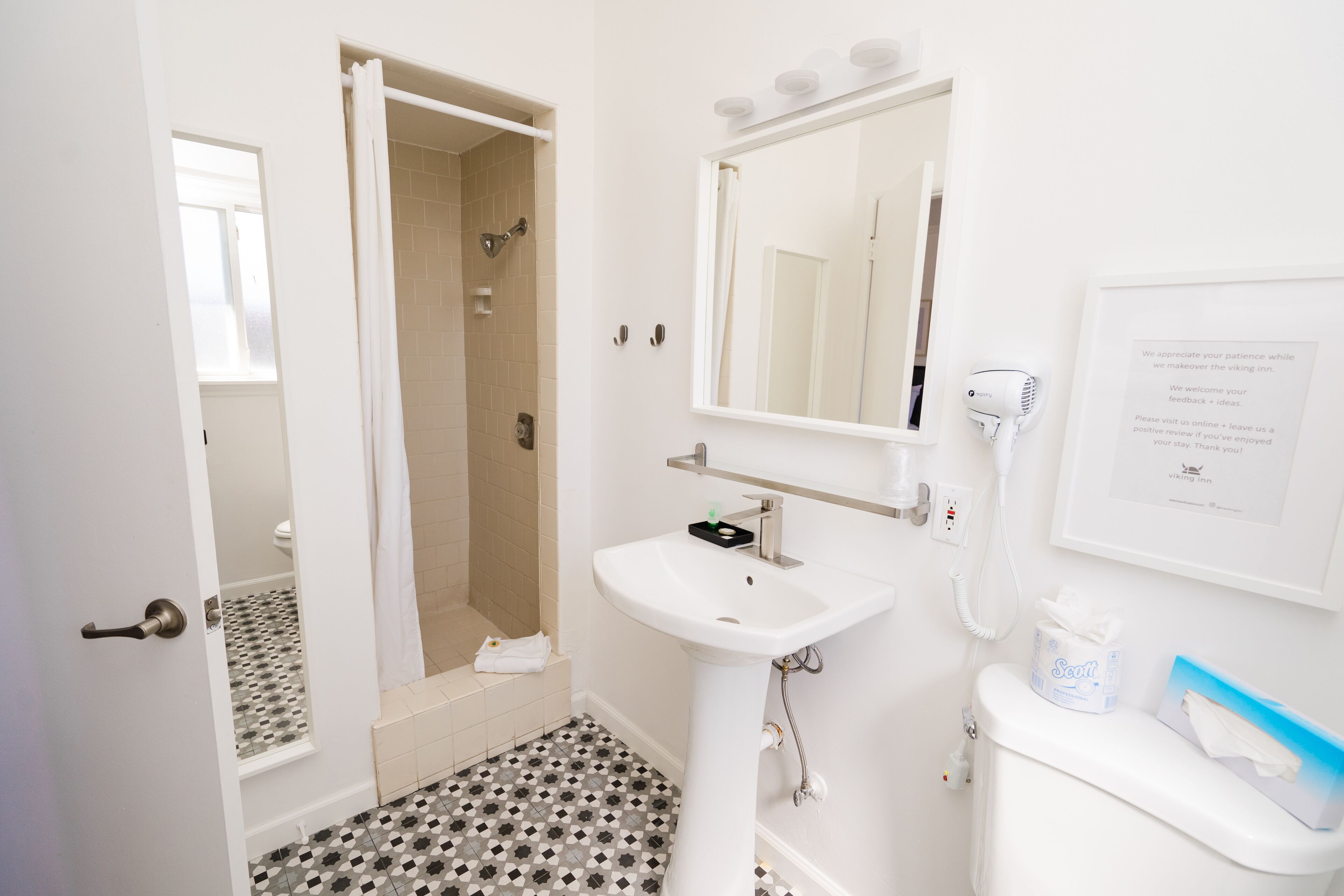 Ein Badezimmer mit Waschbecken, Spiegel und Toilette. Ein Duschvorhang, ein Handtuch und ein gerahmtes Schild sind an der Wand angebracht.